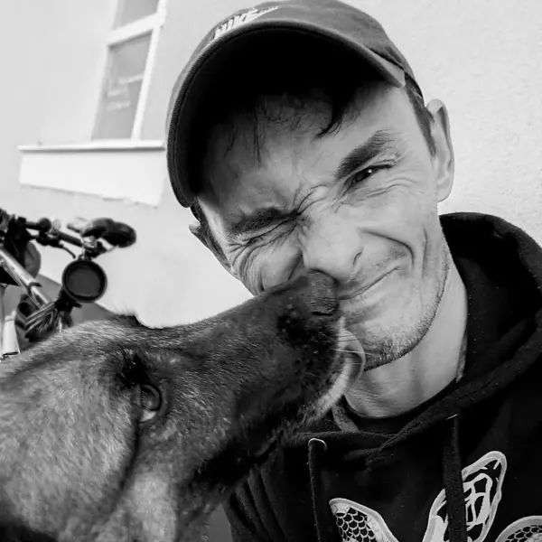 Black and white: a grinning man in a flat cap leans in while a Malinois licks his cheek.